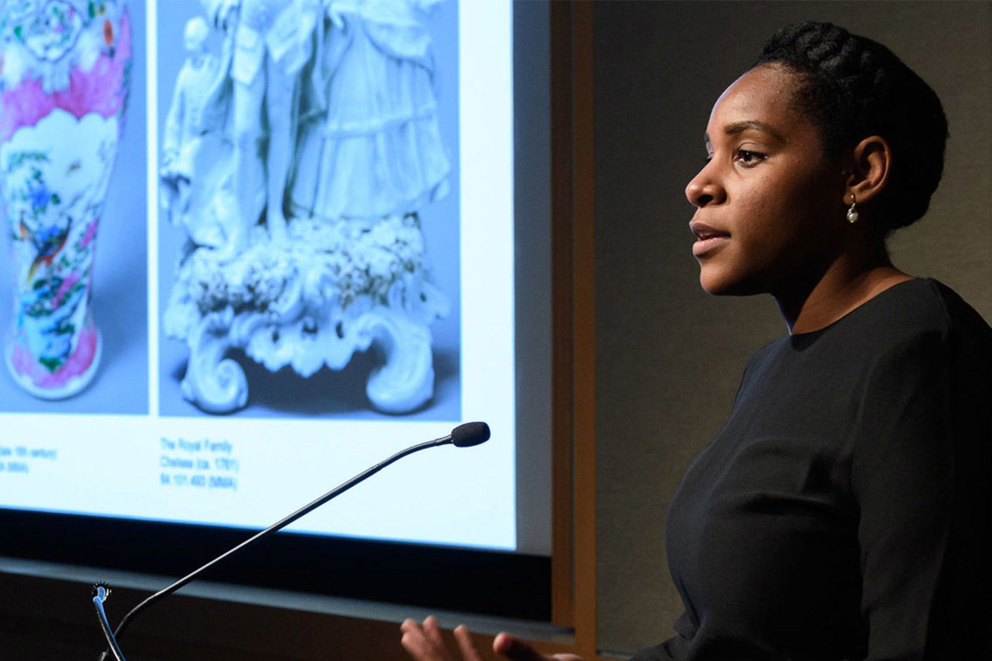 A young woman speaks confidently into a podium microphone in front of a slideshow projection of ceramic artworks